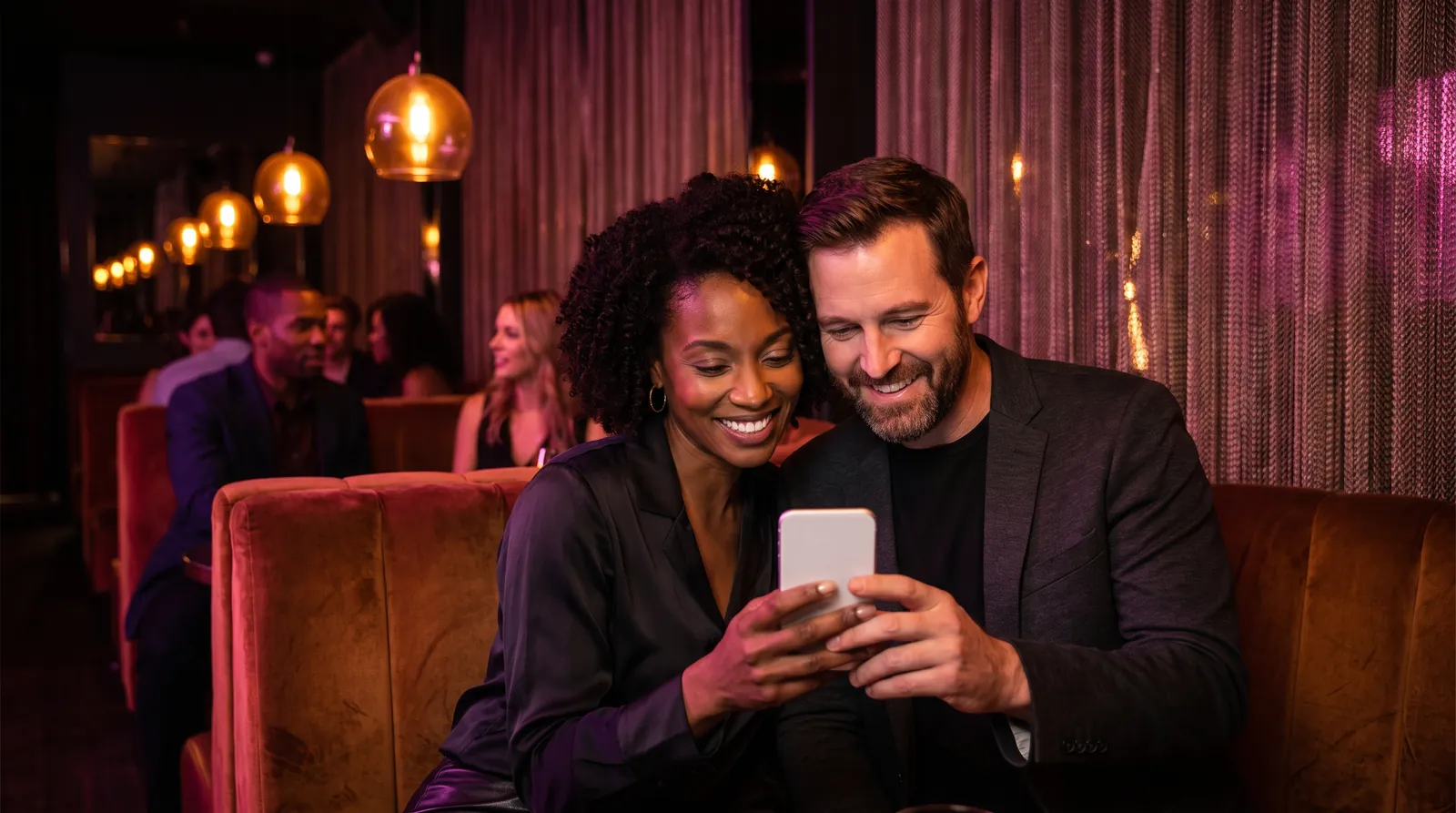 A couple share a smile over a phone in a velvet booth at an upscale lounge