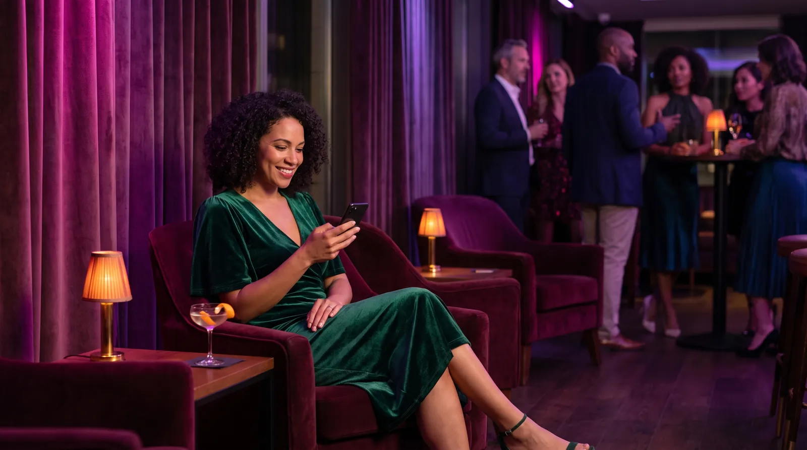 A woman in a green velvet dress smiles at her phone in a plush lounge, cocktail beside her