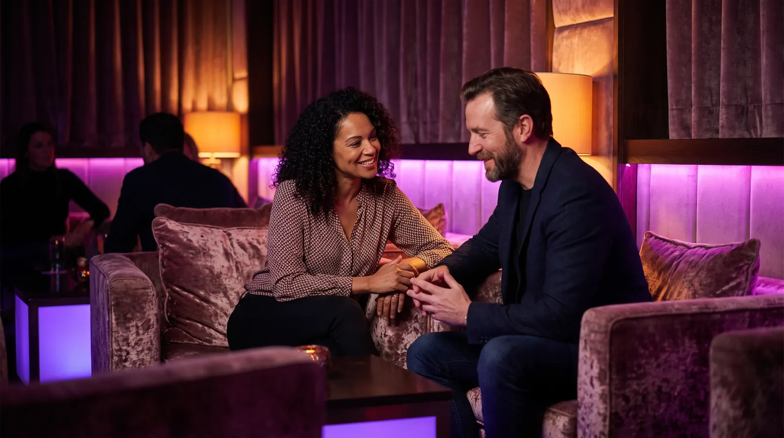 Two adults check in with each other in easy conversation on velvet seating in a warmly lit lounge