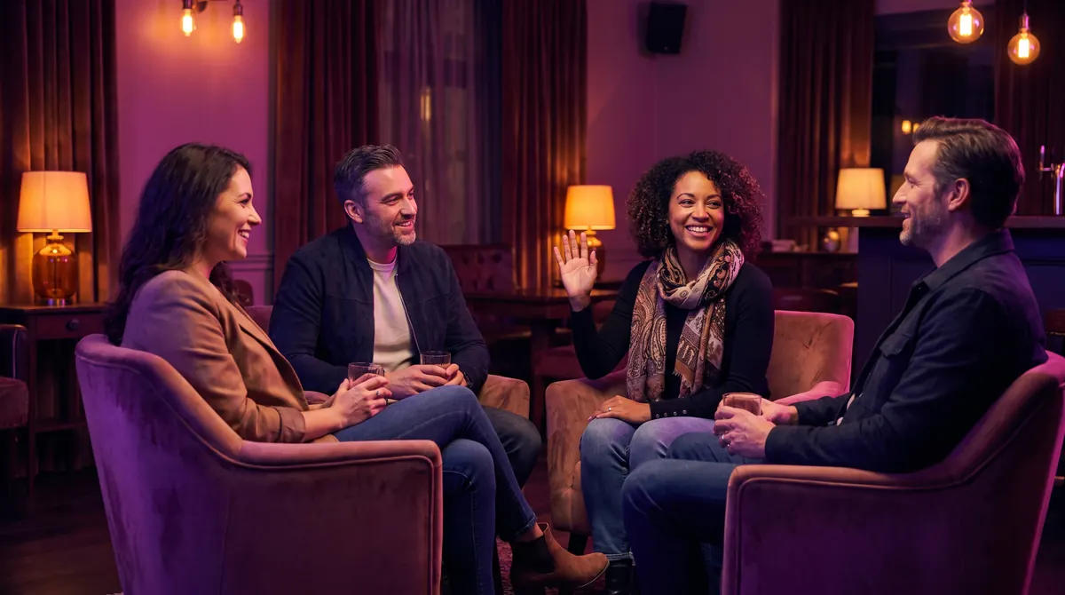 Four friends chat in a warmly lit lounge as one woman smiles and gently raises a hand, the others nodding in easy respect