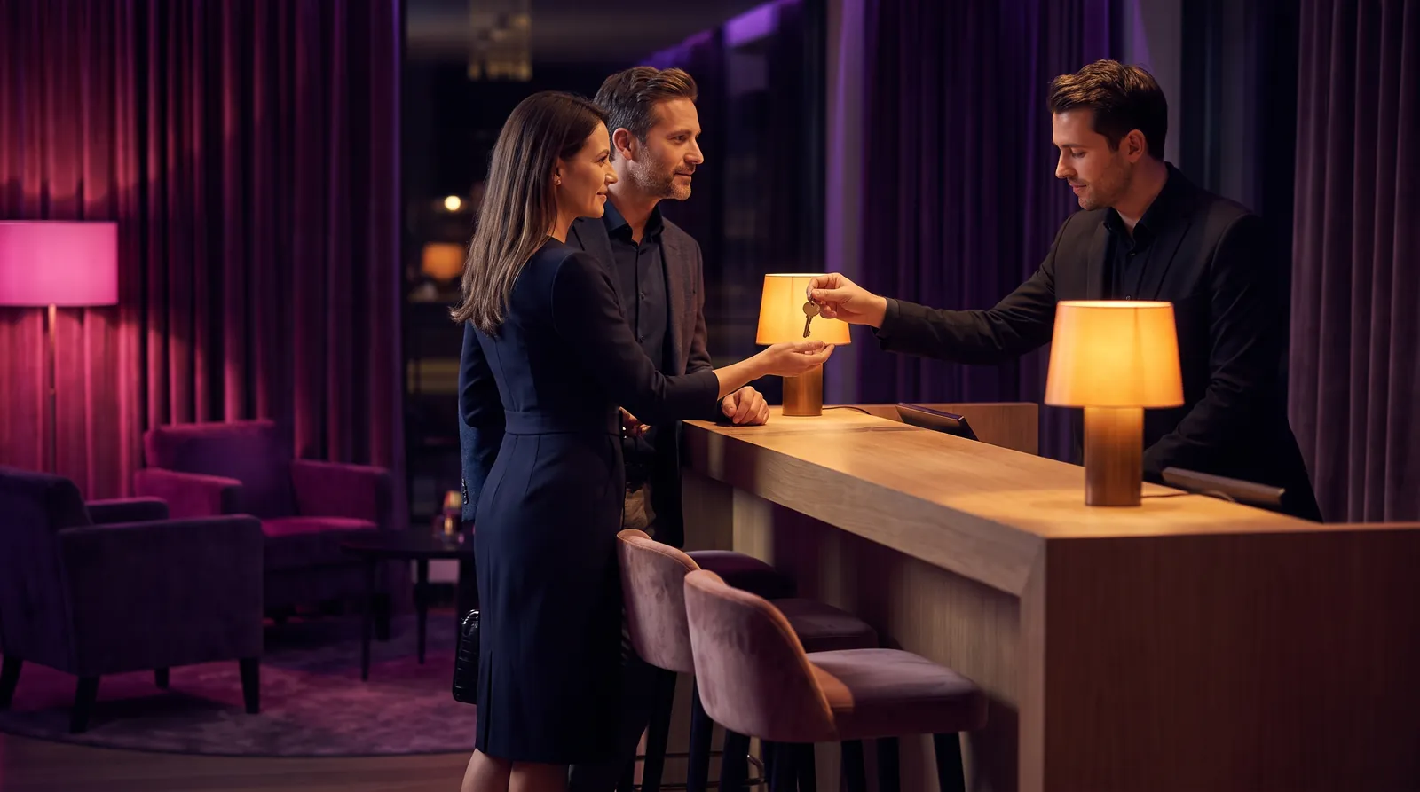 A couple receive a room key from a concierge at a lamplit desk in a velvet lounge