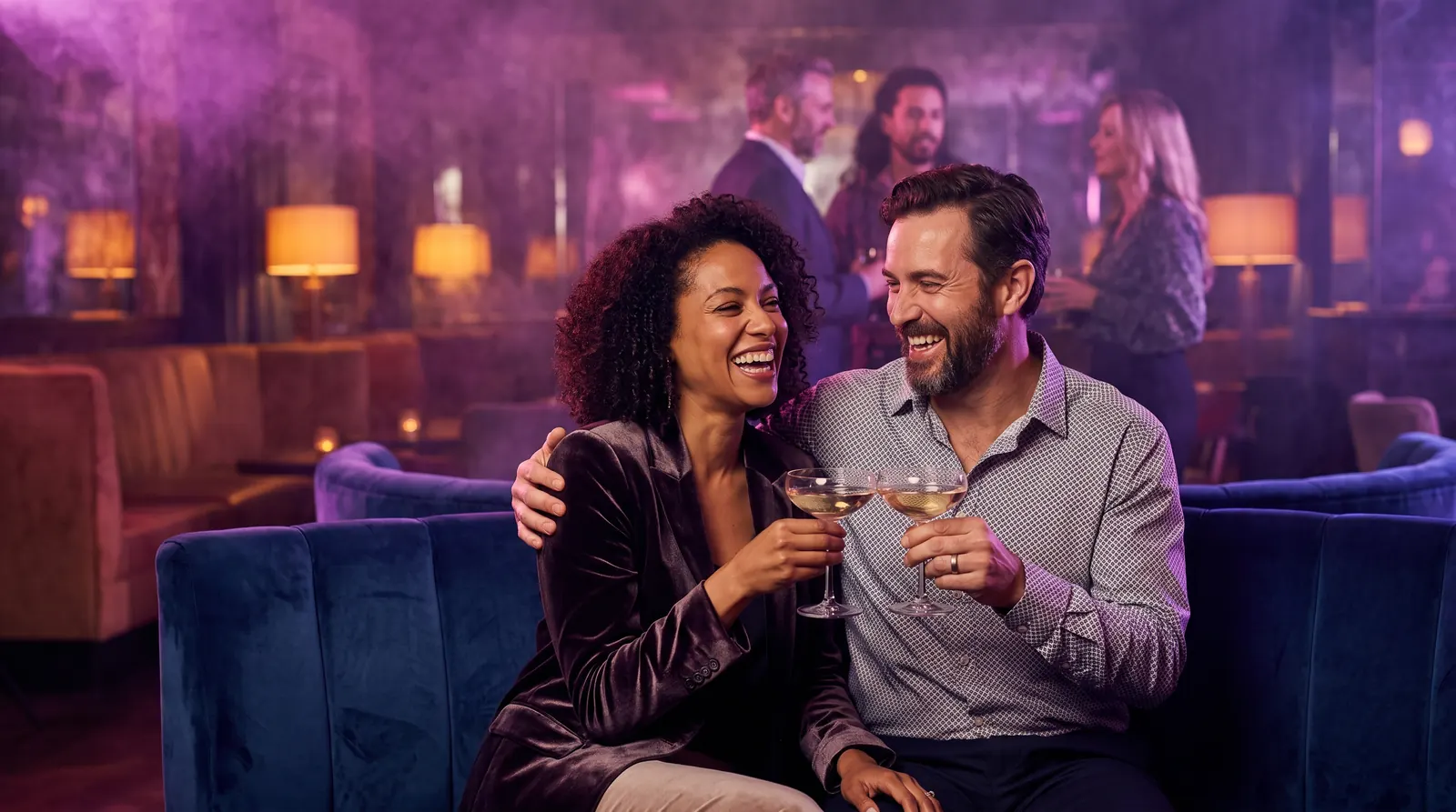 A laughing couple toast with champagne coupes on a blue velvet sofa at a softly glowing lounge party